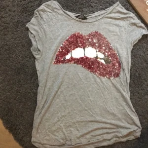 As cool t-shirt - Snygg grå t-shirt med ett stort paljettmotiv av röda läppar på framsidan. Perfekt för att ge din outfit en extra fet touch. T-shirten har korta ärmar och en avslappnad passform.
