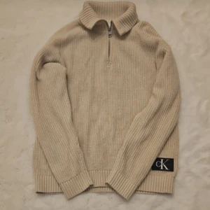 Beige stickad tröja från Calvin Klein Jeans - Säljer en stilren beige stickad tröja från Calvin Klein Jeans med dragkedja vid halsen och ribbade detaljer. Perfekt för kyligare dagar med sin långa ärmar och mjuka material. Ikonisk CK-logga på sidan ger en extra touch av stil.