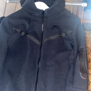 Nike Tech tröja  - Säljer en svart Nike Tech tröja med dragkedja och huva. Tröjan har en stilren design med diskreta detaljer och en ficka på ärmen. Perfekt för en sportig look. Storlek 158-170 vilket motsvarar xxs. Priset kan diskuteras.