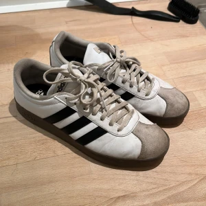 Adidas sneakers i vitt och svart - Säljer ett par klassiska Adidas sneakers i vitt med svarta ränder. Skorna har snörning och en robust sula. Perfekta för en avslappnad stil.