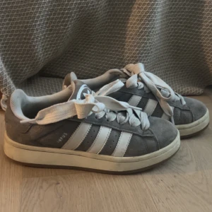 Grå Adidas Campus sneakers - Säljer ett par grå Adidas Campus sneakers i mocka med vita detaljer och snörning. Skorna har den klassiska tre-randiga designen på sidorna och Adidas-loggan på hälen. Perfekta för en avslappnad stil.
