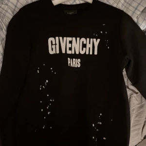 Svart tröja från Givenchy - Säljer en svart tröja från Givenchy med vit text 'GIVENCHY PARIS' på framsidan. Tröjan har långa ärmar och ett stänkmönster i vitt som ger en unik look. Perfekt för dig som vill ha något stilrent men med en twist.