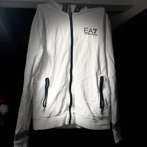 Vit huvtröja från EA7 Emporio Armani - Säljer en stilren vit huvtröja från EA7 Emporio Armani. Tröjan har dragkedja framtill och två praktiska fickor med dragkedjor. Den har långa ärmar och en huva med snygga detaljer. Perfekt för en avslappnad look.