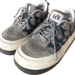 Grå sneakers från Eytys - Säljer ett par grå sneakers från Eytys med unika blå flammor på sidorna. Skorna har en robust design med grå snörning och en vit sula. Det finns en lila detalj på hälen som är lite trasig, 
