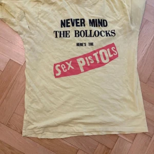 Sex Pistols merch - Cool gul t-shirt med tryck av Sex Pistols album 'Never Mind the Bollocks'. Perfekt för musikälskare och de som gillar retrostil. T-shirten har korta ärmar och en klassisk passform.