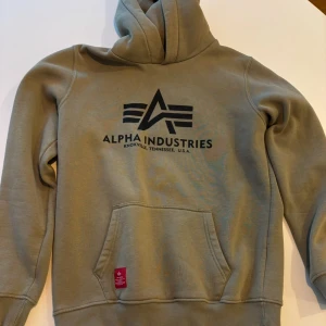 Beige hoodie från Alpha Industries - Snygg beige hoodie från Alpha Industries med svart logga framtill. Tröjan har huva, känguruficka och ribbade muddar vid ärmslut och nederkant. Perfekt för dig som gillar streetwear och vill ha en stilren look.
