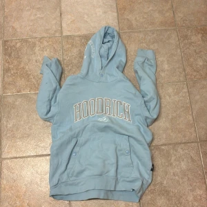 Blå hoodie från Hoodrich - Säljer en ljusblå hoodie från Hoodrich med stor vit logga framtill. Tröjan har huva, känguruficka med knappar och långa ärmar. Perfekt för dig som gillar streetwear och vill ha en chill look.
