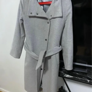 Ljusgrå kappa från Tiger of Sweden - Snygg ljusgrå kappa från Tiger of Sweden med bälte i midjan och knappar framtill. Klassisk och stilren design med lång ärm och bred krage. Perfekt för dig som vill ha en clean och modern look.