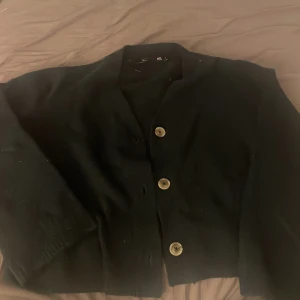 Svart kofta från 157 med knappar - Svart kofta från 157 i storlek M. Klassisk modell med tre stora knappar framtill och lång ärm. Koftan har en rak passform och är tillverkad i ett mjukt stickat material som känns skönt mot huden. Perfekt att slänga över en t-shirt eller topp.