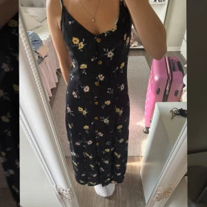 Svart blommig långklänning H&M - Supersöt svart långklänning från H&M med tunna axelband och knappar framtill. Klänningen har ett blommigt mönster i gult och rosa, v-ringad hals och en flowy passform. Perfekt för sommarens alla tillfällen!