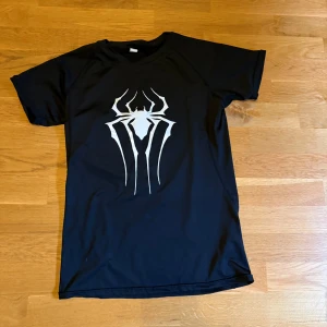 Compression shirt - Cool svart t-shirt med ett stort vitt spindeltryck på bröstet, inspirerat av Spiderman. Klassisk passform med korta ärmar och rund hals. Perfekt för dig som gillar superhjältar och vill sticka ut med en snygg och edgy look.