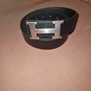 Svart skärp med H-spänne - Säljer ett svart skärp  med klassiskt stort H-spänne i silverfärgad metall. Bältet är gjort i präglat skinn och har en stilren, tidlös look. Perfekt accessoar för att lyfta vilken outfit som helst.