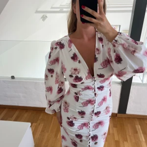 Vit blommig klänning från Gina Tricot - Supersöt vit klänning med rosa och lila blommor från Gina Tricot. Klänningen har ballongärmar, v-ringning och knappar framtill. Figurnära passform och lätt struktur i tyget. Perfekt för sommarens alla tillfällen!