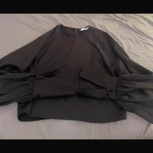 Svart blus med puffärmar från NA-KD - Stilren svart blus från NA-KD med långa puffärmar och dekorativa knappar vid ärmsluten. Blusen har rund halsringning och är tillverkad i ett glansigt, mjukt material som ger en lyxig känsla. Perfekt för dig som gillar trendiga detaljer och elegant look.