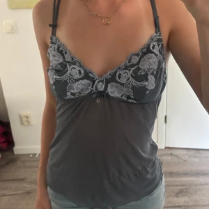 Grå spetslinnetopp med blommor - Supersöt grå linnetopp med tunna axelband och broderade blommor i vitt på bysten. Toppen har en liten rosett och en droppformad detalj framtill. Materialet är tunt och lätt, perfekt för lager-på-lager eller som statement-piece. Ok skick, litet hål under bysten och täcken på användning på sidorna, se bilder🥰