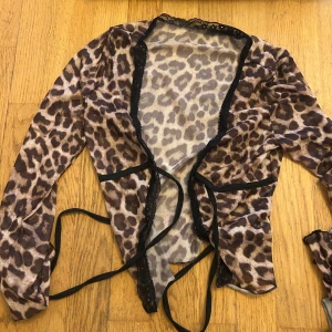 Leopardmönstrad bolerotopp med snörning - Snygg bolerotopp i transparent mesh med leopardmönster och svarta kanter. Toppen har långa ärmar, knytband framtill och spetsdetaljer längs öppningen. Perfekt för att lyfta vilken outfit som helst.