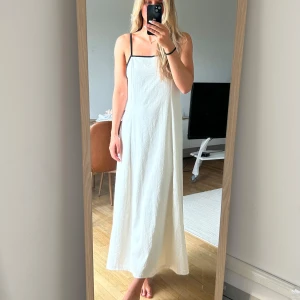 Vit långklänning med svarta band - Superfin vit långklänning med tunna svarta axelband och svart kant längs urringningen. Klänningen har en rak, luftig passform och knytband i ryggen. Perfekt för varma dagar och har en stilren, minimalistisk look.