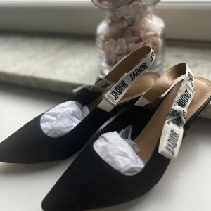 Svarta Dior J'Adior pumps med rosett - Supersnygga svarta pumps från Dior med spetsig tå och vit rem med J'Adior-tryck och rosett. Skorna har låg klack och är tillverkade i textil och skinn. Perfekta för dig som vill ha en lyxig och trendig look. Ny pris 11000kr