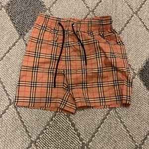 Rutig Burberry badshorts med snörning - Snygga badshorts från Burberry i klassiskt beige och svart rutigt mönster med röda detaljer. Shortsen har elastisk midja med svart snörning och vit mesh på insidan för extra komfort. Perfekta för sommaren och strandhäng.