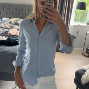 Ljusblå klassisk skjorta i bomull - Säljer en stilren ljusblå skjorta med klassisk krage och knäppning framtill. Skjortan har lång ärm och är figursydd för en snygg passform. Perfekt till jeans eller kostymbyxor.