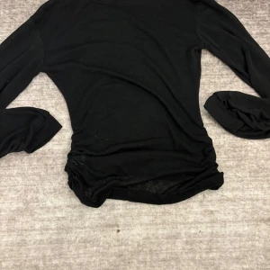 Svart långärmad topp med rynkade sidor - Svart långärmad topp i tunn, lätt transparent bomullsmix. Snygga rynkade detaljer i sidorna ger en cool och trendig look. Perfekt att styla med jeans eller kjol för en enkel men stilren outfit.