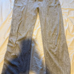 Grå mjukisbyxor Juicy Couture XL - Grå mjukisbyxor från Juicy Couture i storlek XL. Byxorna har resår i midjan, två fickor fram och bakfickor med knappar. Mjukt material som känns skönt mot huden, perfekt för chill dagar. Klassisk Juicy Couture-vibe.