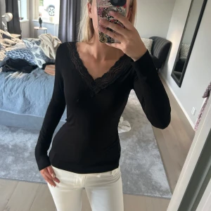 Svart långärmad topp med spets - Säljer en svart långärmad topp med v-ringning och snygga spetsdetaljer längs halsringningen. Modellen är tight och sitter riktigt snyggt på kroppen. Perfekt att styla med både jeans och kjol för en clean look.