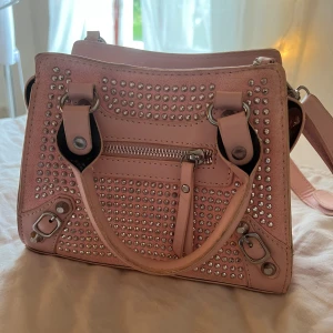 Rosa axelväska med nitar och dragkedja - Snygg liten ljusrosa axelväska med silvriga nitar på framsidan och detaljer i metall. Väskan har både handtag och lång axelrem samt en praktisk dragkedja framtill. Tillverkad i syntetmaterial och har en cool, glittrig vibe. Skriv om du vill ha fler bilder eller ngt annat du undrar över.😊💓
