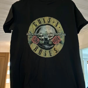 Guns N' Roses svart t-shirt - Svart t-shirt med klassiskt Guns N' Roses-tryck på bröstet. T-shirten har rund hals, korta ärmar och är gjord i mjuk bomull. Perfekt för dig som älskar rock och vill ha en ikonisk bandtröja i garderoben.