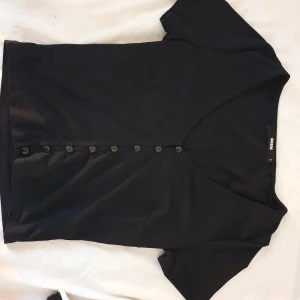 Svart topp med knappar från Bik Bok - En svart kortärmad topp från Bik Bok med v-ringning och knappar hela vägen framtill. Snygg och enkel design som passar till mycket. Gjord i mjukt material och har en figurnära passform.