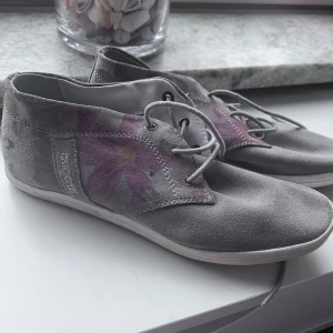 Grå mockasneakers med blommigt mönster - Unika sneakers från Biogateo i grå mocka med snyggt blommigt mönster i lila och gult på sidorna. Skorna har snörning och vit platt sula, perfekt för dig som vill ha något annorlunda och stilrent. Storlek 36.