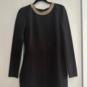 Svart klänning med gulddetaljer - Supersnygg svart kort klänning med långa ärmar och öppen rygg. Runt halsen och längs ryggen finns coola guldfärgade pärldetaljer som ger en lyxig känsla. Klänningen har en figurnära passform och är perfekt för dig som vill sticka ut