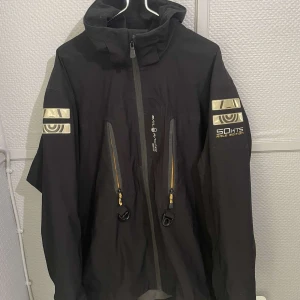 Svart Gore-Tex vindjacka Sail Racing XL - Svart vindjacka från Sail Racing i Gore-Tex-material. Jackan har huva, två snedställda dragkedjefickor framtill och coola reflexdetaljer på ärmarna. Broderad logga på bröstet och tryck på ärmen. Perfekt för dig som gillar stilren och funktionell design.