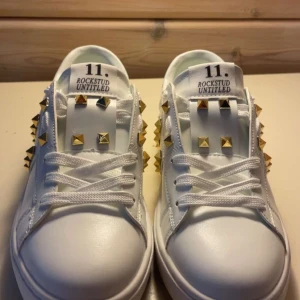 Valentino Garavani Rockstud sneakers - Snygga vita Valentino Garavani Rockstud sneakers i skinn med ikoniska guldiga nitar på sidorna och tungan. Klassisk rund tå, platt sula och coola detaljer längs hälen. Perfekta för dig som vill sticka ut med lyxig streetstyle.