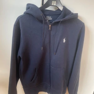 Marinblå zip-hoodie från Polo Ralph Lauren - Snygg marinblå zip-hoodie från Polo Ralph Lauren med dragkedja och klassisk vit logga på bröstet. Tröjan har huva med snörning, två fickor fram och är tillverkad i mjuk bomull. 