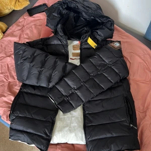 Svart dunjacka från Parajumpers - Säljer en svart dunjacka från Parajumpers med klassisk patch på ärmen och gul detalj vid kragen. Jackan har huva, dragkedja och är fodrad i vitt. Perfekt för kalla dagar och riktigt snygg streetstil.