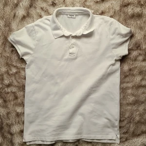 Vit pikétröja från Kappahl - Klassisk vit pikétröja från Kappahl i bomull. Tröjan har korta ärmar, krage och knäppning med tre knappar framtill. Perfekt basic-plagg med stilren look och mjukt material som passar till många outfits.