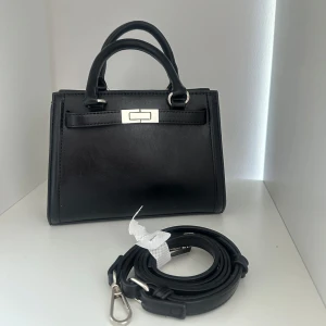 Svart handväska med axelrem - Stilren svart handväska i skinnimitation med silverfärgade metalldetaljer och avtagbar axelrem. Väskan har två handtag och ett klassiskt spänne framtill. Perfekt storlek för det viktigaste och passar till många olika looks.