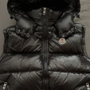 Svart Moncler dunväst med huva - Snygg svart dunväst från Moncler med glansig finish och ikonisk logga på bröstet. Västen har en rejäl huva, dragkedja framtill och coola detaljer som tryckknappar vid axlarna. Perfekt för lager-på-lager och streetstyle.