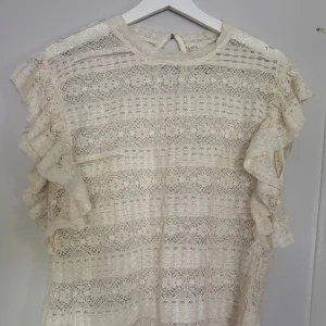 Beige spetsblus med volangärmar - Superfin beige blus i spets med volangärmar och rund halsringning. Blusen har ett vackert spetsmönster över hela och knäppning bak i nacken. 