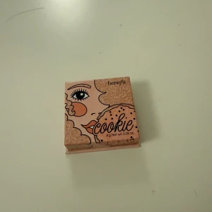 Benefit Cookie highlighter - Supersnygg highlighter från Benefit i nyansen Cookie. Kommer i en söt rosa ask med spegel i locket. Ger en intensiv champagneguldig glow som verkligen poppar på kindbenen. Perfekt för dig som älskar att glänsa och vill ha en ikonisk look.
