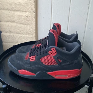 Nike Air Jordan 4 svart/röd sneakers - Säljer ett par Nike Air Jordan 4 sneakers i svart mocka med röda detaljer och meshpaneler. Skorna har klassisk Jumpman-logga på hälen och plös, samt svart snörning. Snygg kontrast mellan svart och rött gör dem till ett statement i varje outfit.