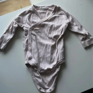 Randig omlottbody från Newbie, stl 68 barn - Supersöt omlottbody från Newbie i storlek 68. Ljusrosa och vita tunna ränder, långärmad och med praktiska tryckknappar längs sidan och i grenen. Tillverkad i mjuk bomull, perfekt för småttingar.