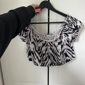 Zebramönstrad croppad topp från Gina Tricot - Säljer en croppad topp från Gina Tricot med svartvitt zebramönster. Toppen har korta puffärmar och resår vid halsringningen som ger en söt volangkant. Perfekt för dig som vill sticka ut med ett djurmönster och gillar trendiga detaljer.
