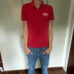Röd Peak Performance pikétröja - Snygg röd pikétröja från Peak Performance med klassisk krage och knappar framtill. Tröjan har korta ärmar och vit broderad logga på bröstet. Tillverkad i mjuk bomull som känns skön mot huden. Perfekt för en clean och sportig stil.