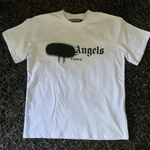 Palm Angels t-shirt - Vit t-shirt från Palm Angels med ikoniskt svart spray paint-tryck och texten 'Angels Paris' på bröstet. Klassisk passform med rund hals och korta ärmar. Tillverkad i mjuk bomull, perfekt för en clean streetwear-look. ENBART TESTAD EN GÅNG. Helt ny! 