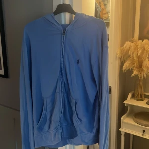 Blå hoodie från Polo Ralph Lauren - Blå hoodie från Polo Ralph Lauren med dragkedja framtill och klassisk broderad logga på bröstet. Tröjan har huva, kängurufickor och är tillverkad i mjuk bomull. Perfekt för en chill och stilren look.