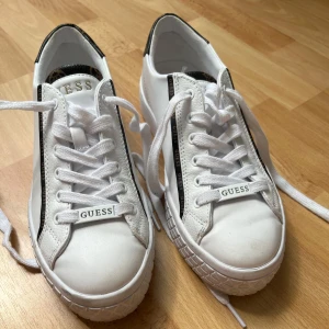 Vita sneakers från Guess med logga - Snygga vita sneakers från Guess med svarta och bruna detaljer samt logotyp längs sidorna och på hälen. Skorna har snörning, platt sula och rund tå. Materialet är syntet med inslag av skinnimitation. Perfekta för en trendig och clean look.