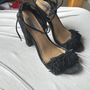 Svarta mockasandaler Steve Madden - Svarta sandaler från Steve Madden i mocka med höga klackar och öppen tå. Skorna har snörning runt vristen och coola fransdetaljer framtill. Perfekta för dig som vill sticka ut med en trendig och edgy look.