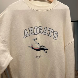 Vit sweatshirt Axel Arigato med print - Snygg vit sweatshirt från Axel Arigato med stort tryck framtill av ett flygplan och texten 'ARIGATO'. Tröjan har rund hals, ribbade muddar och en relaxed passform. Använd 1 gång. Storlek L. Nypris 2599kr